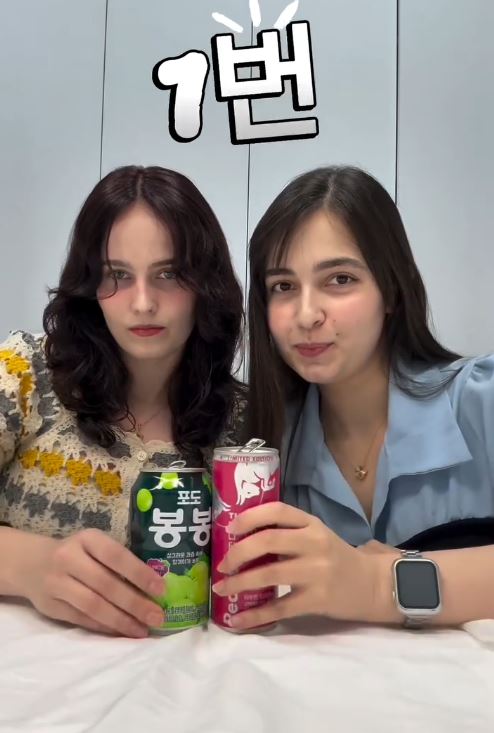 Two young women posing with canned beverages, one green and one pink, on a table. The background is minimalistic with soft lighting.