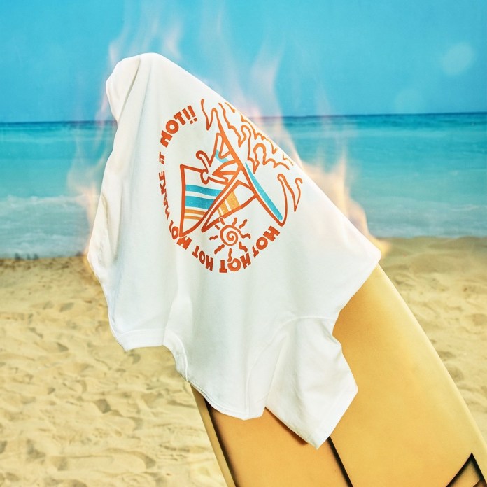 A white t-shirt draped over a surfboard, featuring a colorful graphic that says 'Make it HOT!!' with surf and sun motifs, set against a beach background.