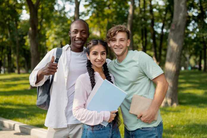 Three young adults smiling together in a park, two holding books and one giving a thumbs up.