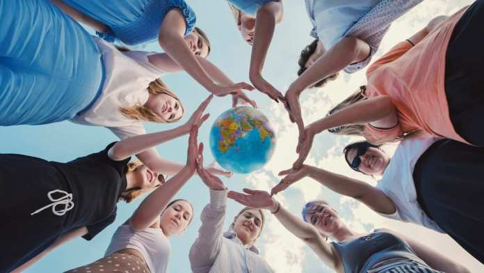 A group of diverse women gathered in a circle, placing their hands together around a globe, symbolizing unity and global connection against a clear blue sky.