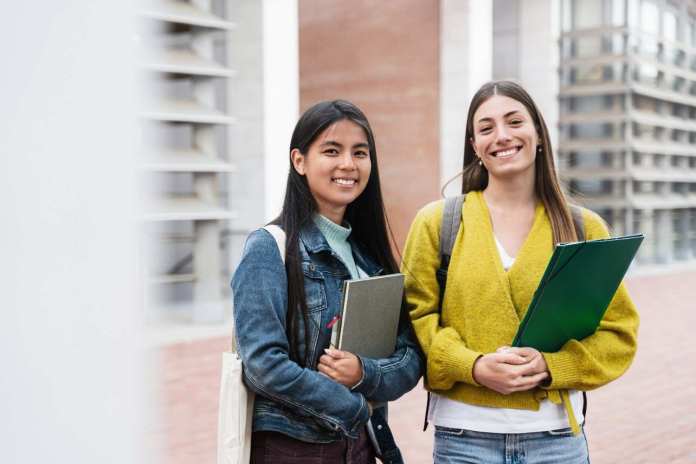 Two students smiling together outdoors, one wearing a denim jacket with notebooks, and the other in a yellow sweater holding folders.