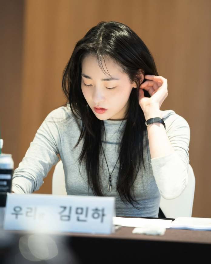 A focused woman with long black hair, wearing a gray long-sleeve shirt, sits at a table while touching her hair and looking down at documents.