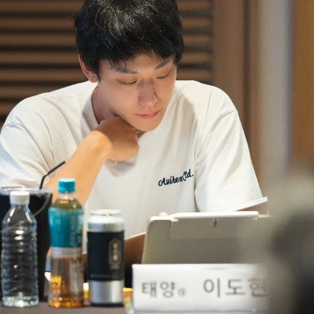 A young man in a white t-shirt is focused on reading or writing at a table, surrounded by water bottles and a laptop, with a nameplate in front of him.