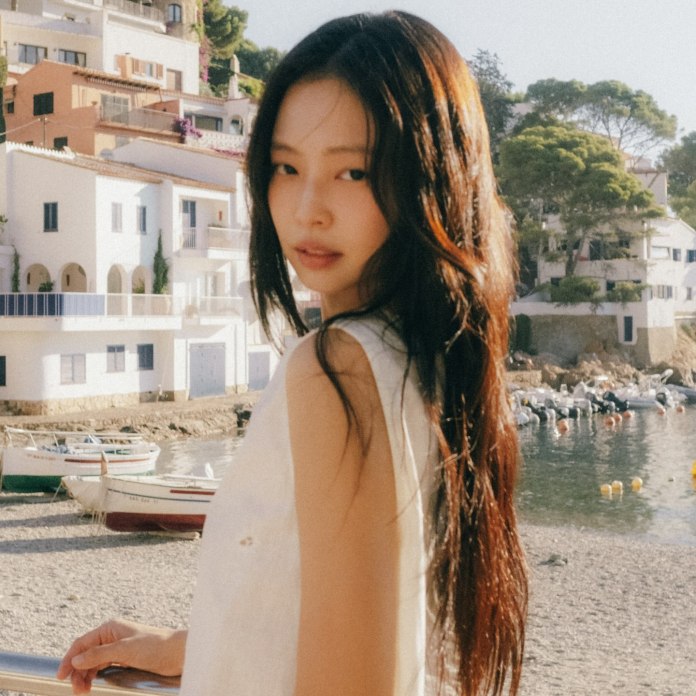 Jennie with long hair, wearing a sleeveless top, gazes over her shoulder at the camera. Behind her, there are white buildings and boats along a seaside harbor.
