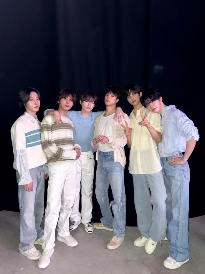 NCT DREAM members posing together in front of a black backdrop, wearing fashionable casual outfits. They are standing close, with one person making a peace sign.