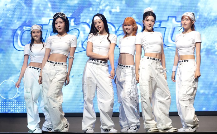 AtHeart performing on stage, dressed in matching white crop tops and baggy white pants with the text 'ATHEART' on the waist. They are posing confidently against a blue background.
