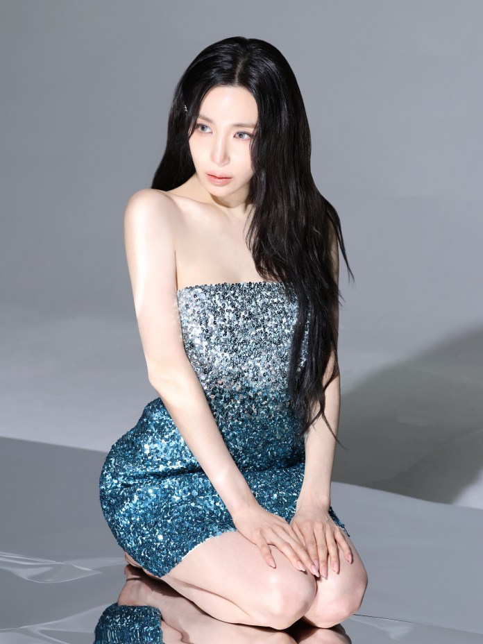 Tiffany Young seated on a reflective surface, wearing a strapless sequined dress in shades of blue and silver. The background is neutral and the lighting highlights their face and outfit.