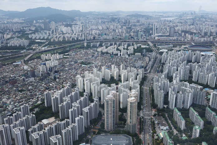 Aerial view of a densely populated urban area featuring numerous high-rise buildings, interspersed with smaller structures and green spaces, set against a backdrop of mountains and a cloudy sky.