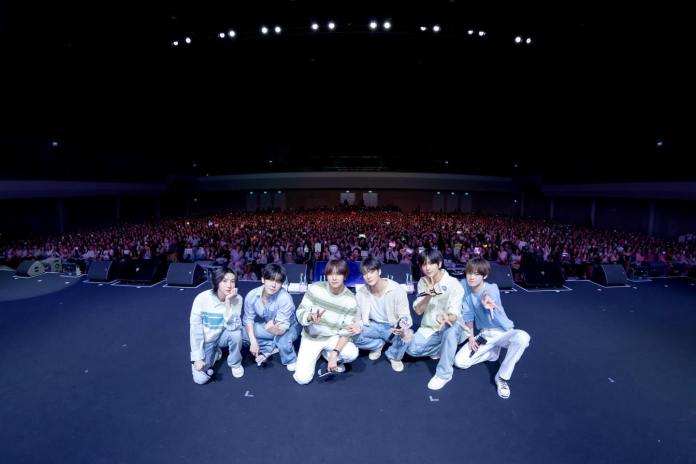 NCT DREAM members pose in front of a large audience at a concert, with a sea of fans in the background holding signs and lights.