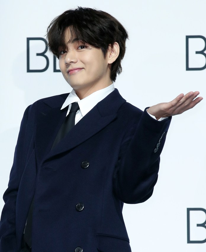 V in a dark suit with a white shirt and black tie smiles and gestures with his hand, posing at an event.