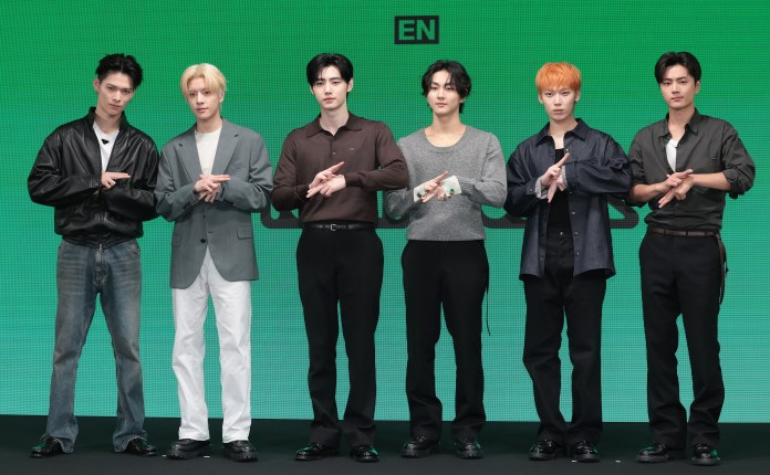 ENHYPEN  members standing in a row, posing for a photo with their hands positioned in a unique gesture. They are dressed in stylish outfits, featuring a mix of casual and formal attire, with a green background behind them.