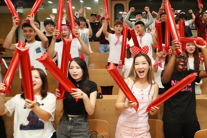 A diverse group of young people enthusiastically cheering and celebrating, holding up large red inflatable cheering sticks in a lively setting.