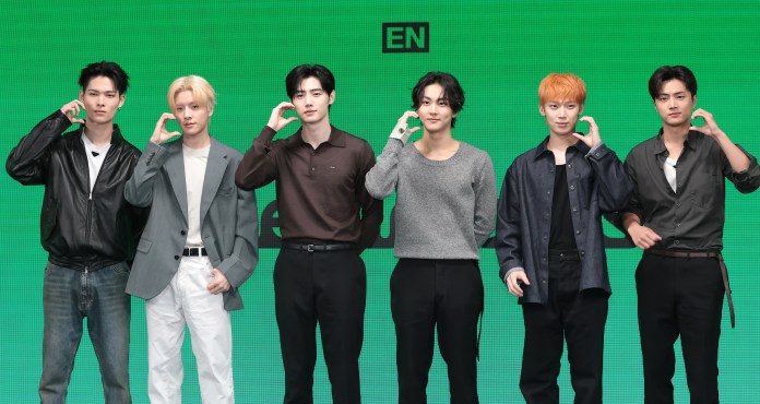 ENHYPEN members posing during a promotional event, each making a hand gesture by placing their hand to their ear, with a green background.