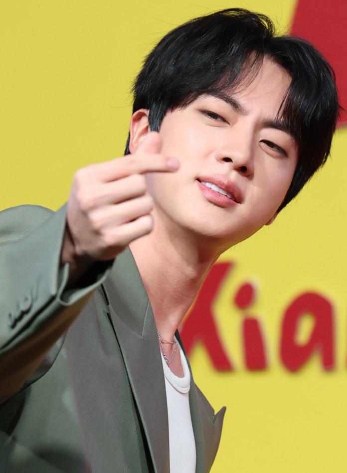 Jin with dark hair, wearing a stylish jacket, making a hand gesture toward the camera against a bright yellow background.