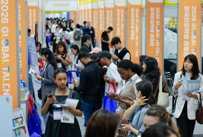 A busy career fair scene featuring diverse attendees engaging in discussions, viewing information materials, and using mobile devices, with banners for the '2026 Global Talent' event in the background.