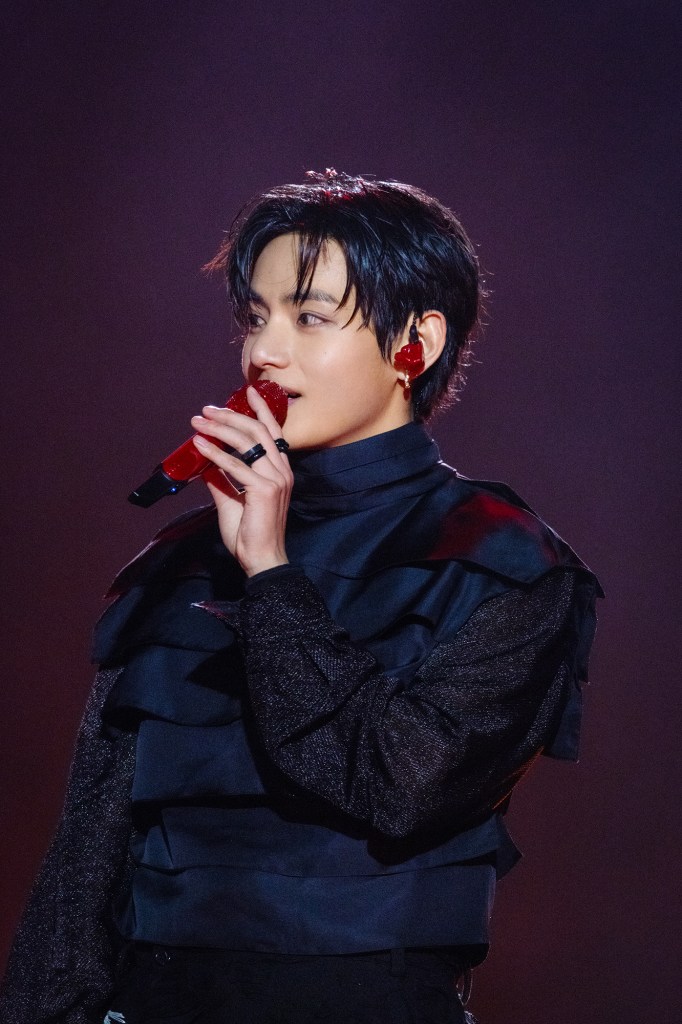 V singing into a red microphone, wearing a stylish black outfit with ruffled details, against a dark background.