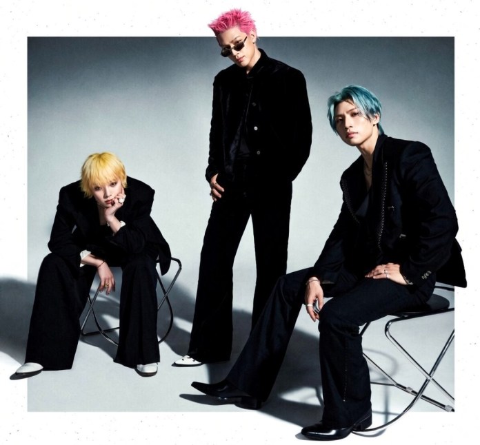 BIGBANG posing in stylish black outfits with colorful hair: one with pink hair standing, one with yellow hair sitting, and one with blue hair sitting.