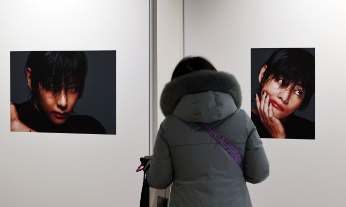 A person in a gray coat is viewing two artistic portraits displayed on a wall, featuring close-up images of V with expressive facial features.