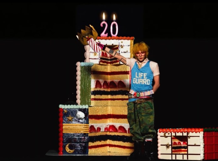 A performer wearing a lifeguard tank top and green camouflage pants stands beside a colorful, large cake adorned with candles and decorations, celebrating a birthday with the number '20' on top.