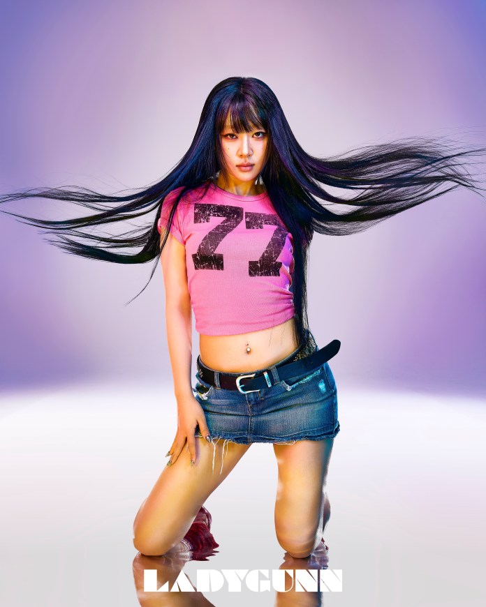 Yves with long black hair poses confidently, wearing a pink shirt with the number 77 and a denim mini skirt. She kneels with one hand on her thigh and the other resting on her knee, against a softly lit purple background.