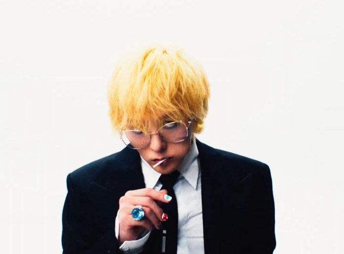Person with blonde hair wearing glasses and a black suit, holding a lollipop and looking down against a white background.