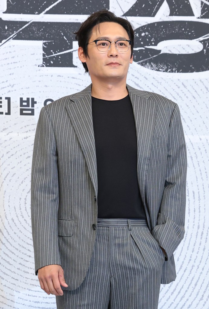 A man in a striped suit and glasses stands confidently, with a neutral expression, against a textured background.