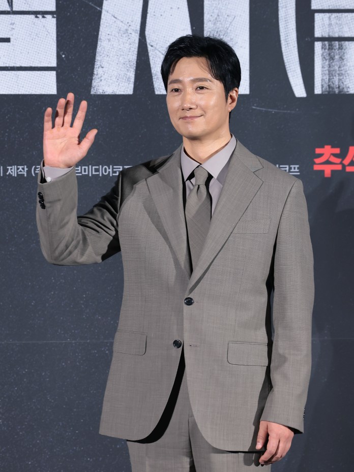 Park Hae-il in a light gray suit with a tie waves at the camera during an event.