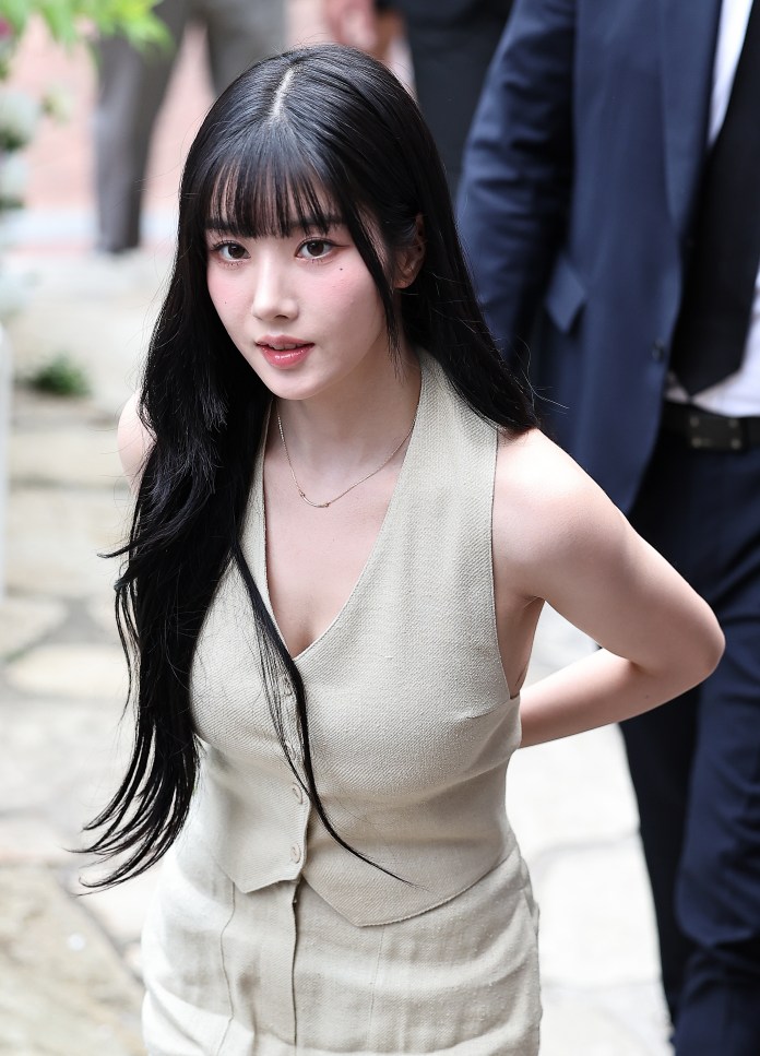 Kwon Eun-bi with long black hair and bangs, wearing a light-colored sleeveless dress, walking outdoors, with a group of people in the background.