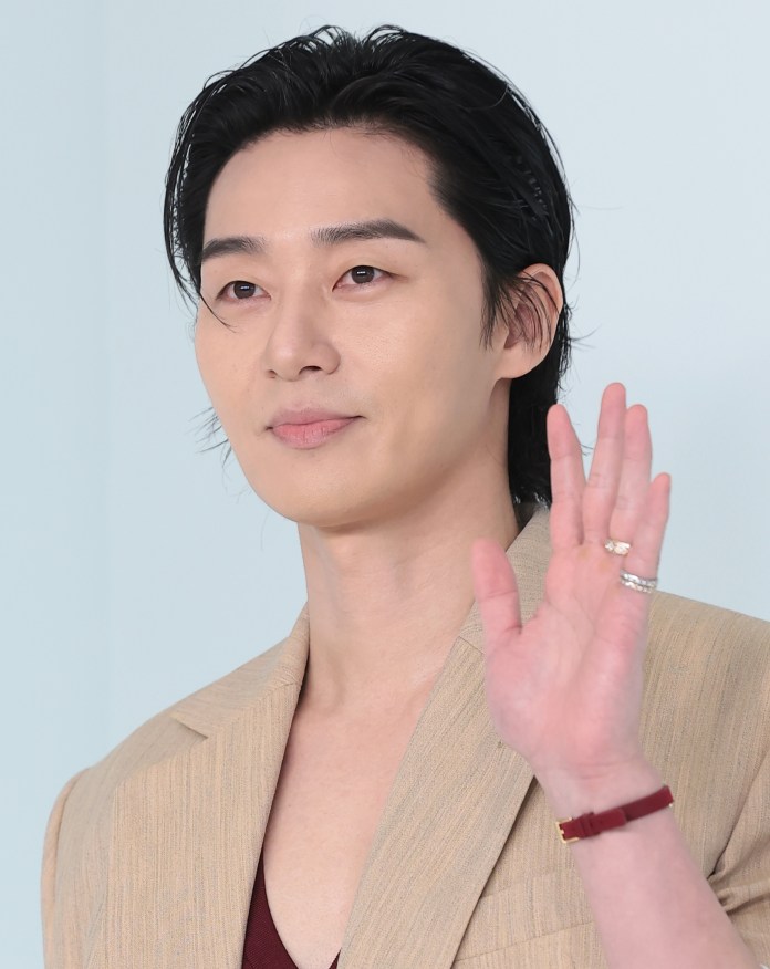 Park Seo-joon with stylish hair is raising his hand, wearing a beige blazer over a burgundy shirt.