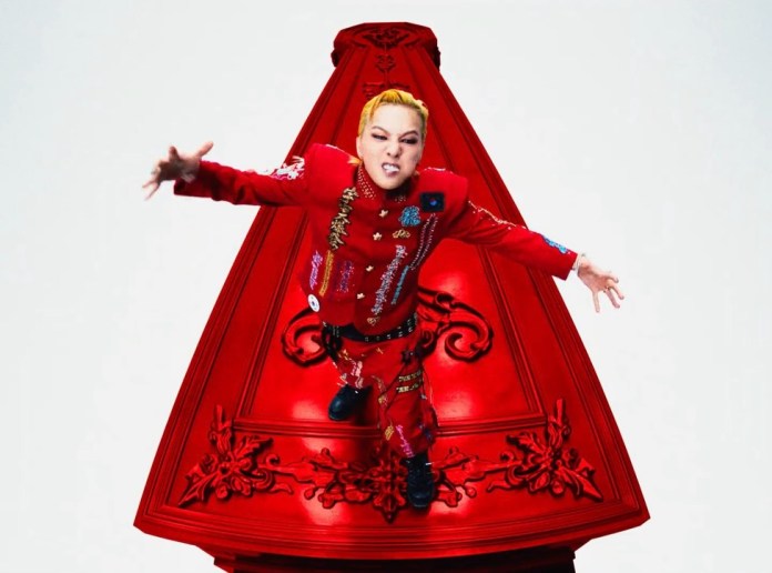 Person in a red outfit with unique embellishments, standing on a red platform with a triangular shape, making an intense expression.