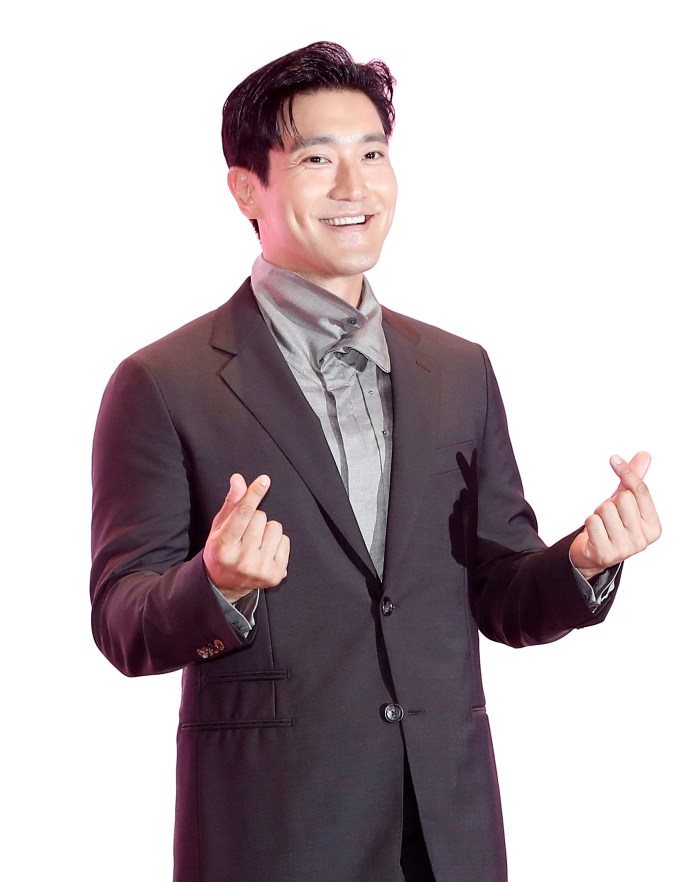 Choi Si-won in a dark suit and grey shirt making a heart gesture with his hands against a white background.