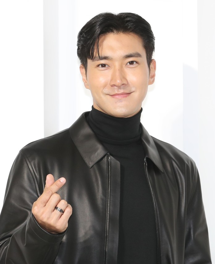 Choi Si-won wearing a black leather jacket and turtleneck smiling and making a hand gesture with their fingers.