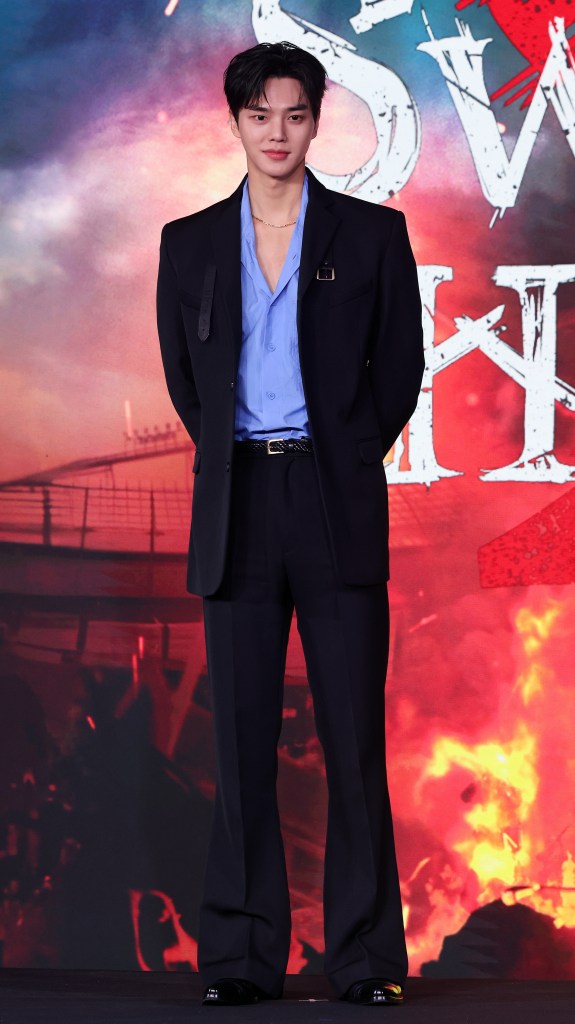 Song Kang in a stylish outfit stands confidently at a promotional event, with a vibrant and dramatic background.
