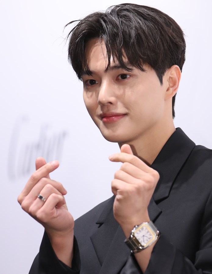 Song Kang with styled dark hair smiles and makes finger heart gestures, wearing a black suit and a silver wristwatch.