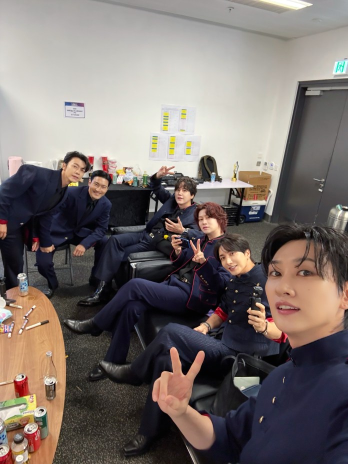A group of six individuals dressed in matching dark outfits, smiling and posing for a selfie in a casual indoor setting. They are seated on a couch with snacks and drinks on a table in front of them.