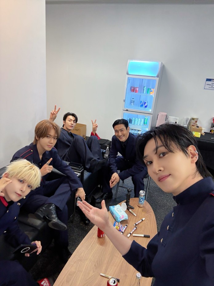 Five individuals in matching navy outfits pose for a selfie in a casual setting, sitting at a table with snacks and drinks. A refrigerator with beverages is visible in the background.