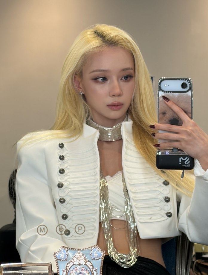 Winter with long blonde hair is seated in front of a mirror, wearing a stylish white military-inspired jacket with decorative buttons and layered necklaces. She is holding a smartphone for a selfie, displaying a fashionable look.