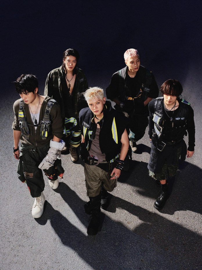 NCT dressed in stylish, casual outfits with utility elements, posing confidently in a dramatic setting. They appear to be standing on an outdoor surface with a dark background.