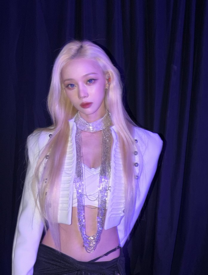 Winter with long blonde hair wearing a stylish white blazer with buttons and a silver necklace, standing in front of a dark backdrop.