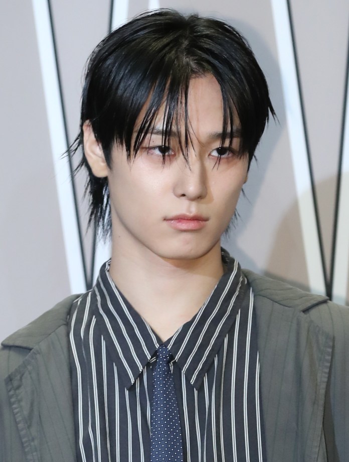 Close-up of Juyeon with black, slicked-back hair, wearing a striped shirt and a tie, looking serious against a neutral background.
