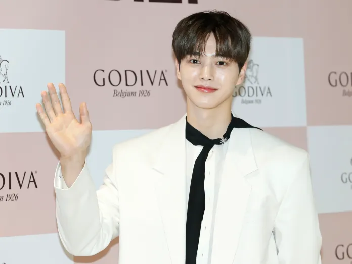 Song Kang in a white suit and black tie waves at the camera at a promotional event with Godiva branding in the background.