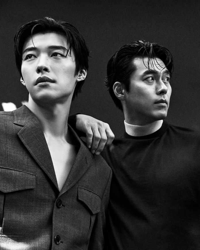 Woo Do-hwan and Hyun Bin pose closely together in a black and white photograph, Woo Do-hwan wearing a gray blazer and Hyun Bin a black shirt. Their expressions are thoughtful, with one man's hand resting on the other's shoulder.