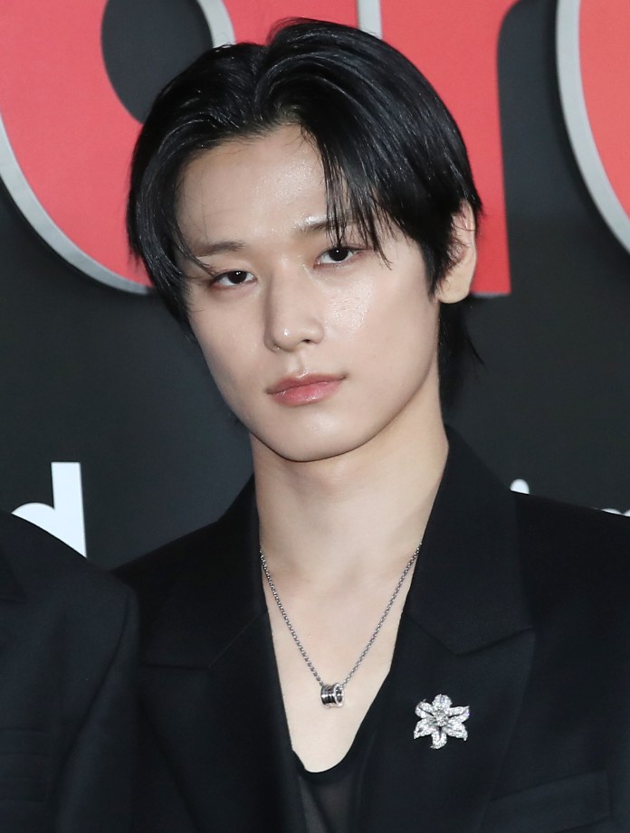 Close-up of Juyeon with dark, shoulder-length hair, wearing a black suit, looking directly at the camera. They have a silver necklace and a flower-shaped brooch on their lapel.