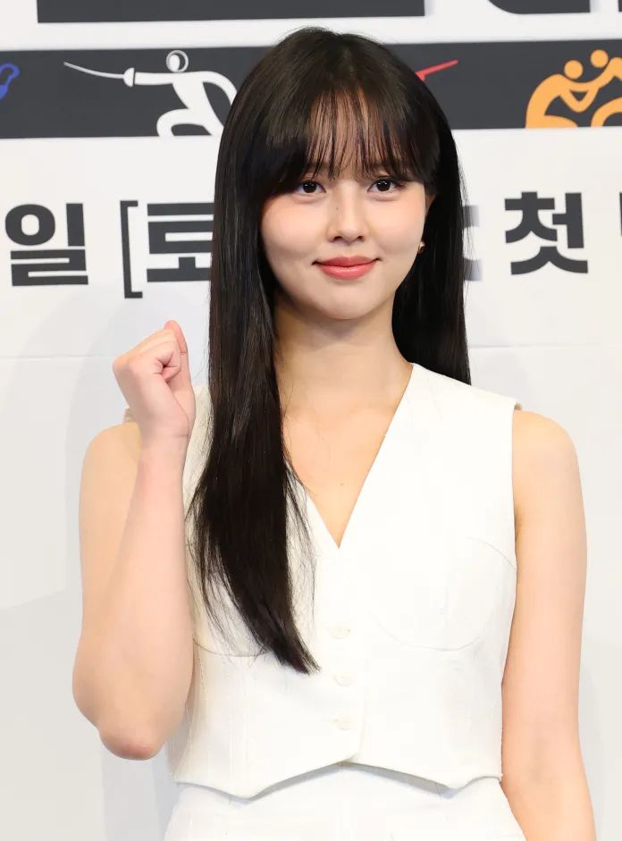 Kim So-hyun wearing a white outfit, smiling and making a fist gesture with her right hand, standing in front of a backdrop with various symbols related to sports and events.