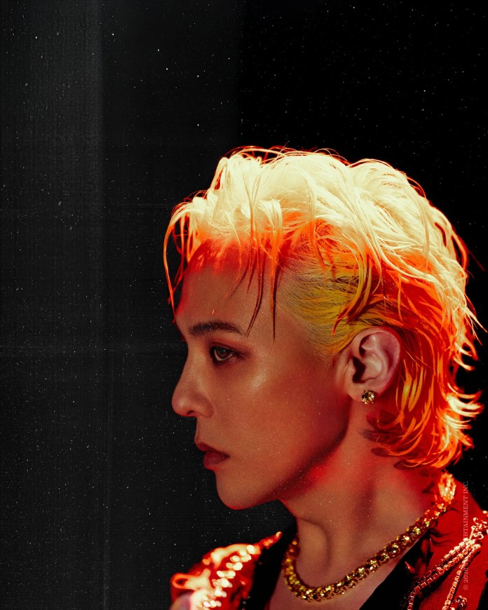 Side profile of G-Dragon with blonde hair and orange highlights, wearing a jeweled necklace and a stylish outfit, against a dark background.