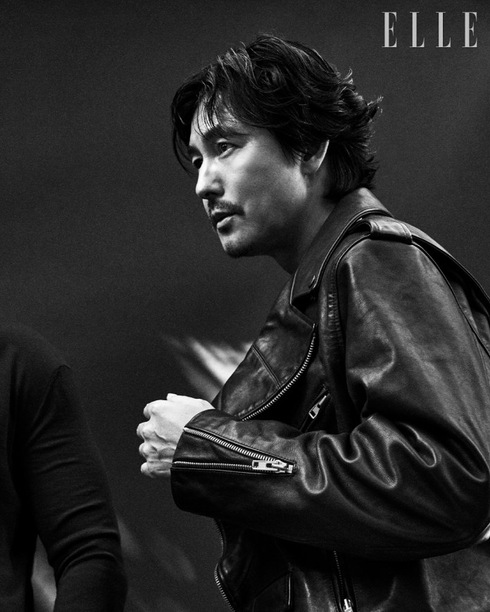 A close-up black-and-white portrait of Jung Woo-sung in a leather jacket, looking thoughtfully to the side, against a dark background. The image captures a sense of intensity and style.