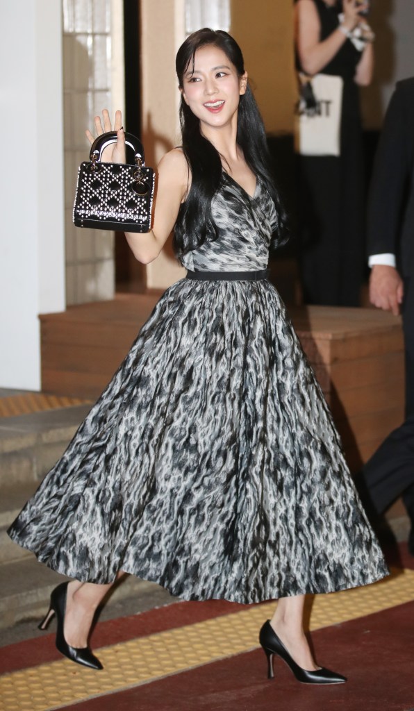 Jisoo in a stylish black and white dress, smiling and waving, holds a small handbag as she steps outdoors.