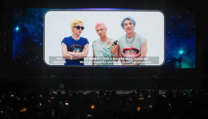 BIGBANG members with colorful hairstyles are featured on a large screen showing a cosmic background, with text in both English and Korean announcing an upcoming event.