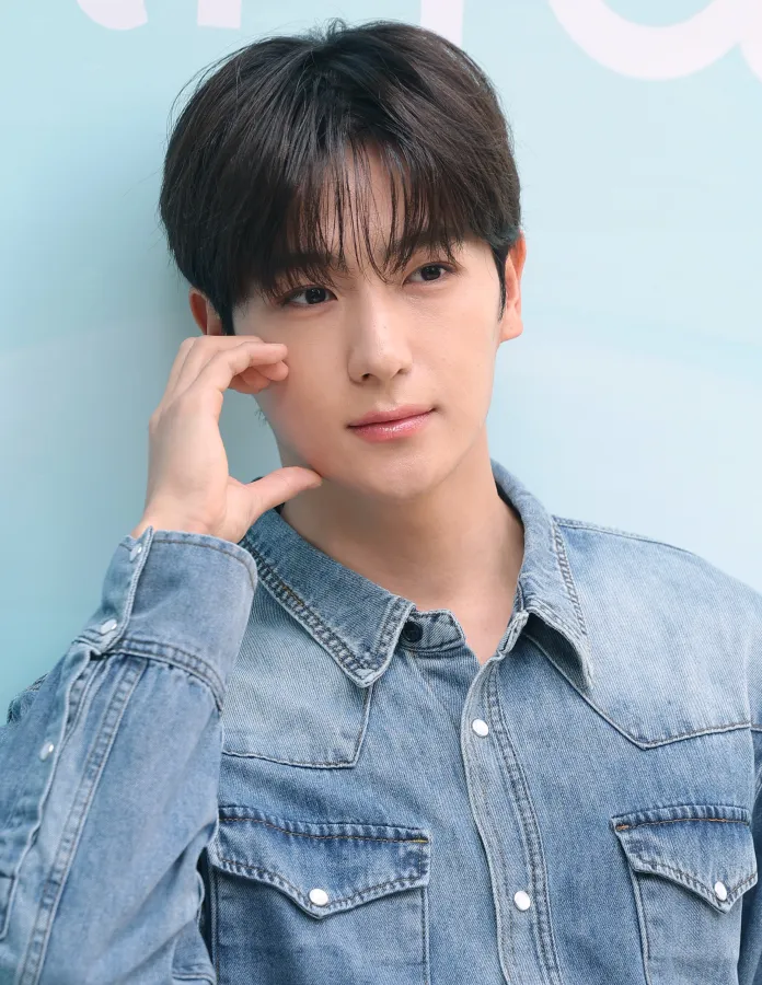Hyunjae with dark, styled hair wearing a light denim jacket, resting his chin on one hand and looking thoughtfully at the camera against a soft blue background.