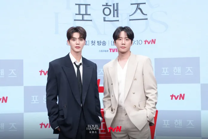 Song Kang and Lee Jun-young  posing together at a promotional event for a TV show, wearing formal attire. The background features the show's title and logo.